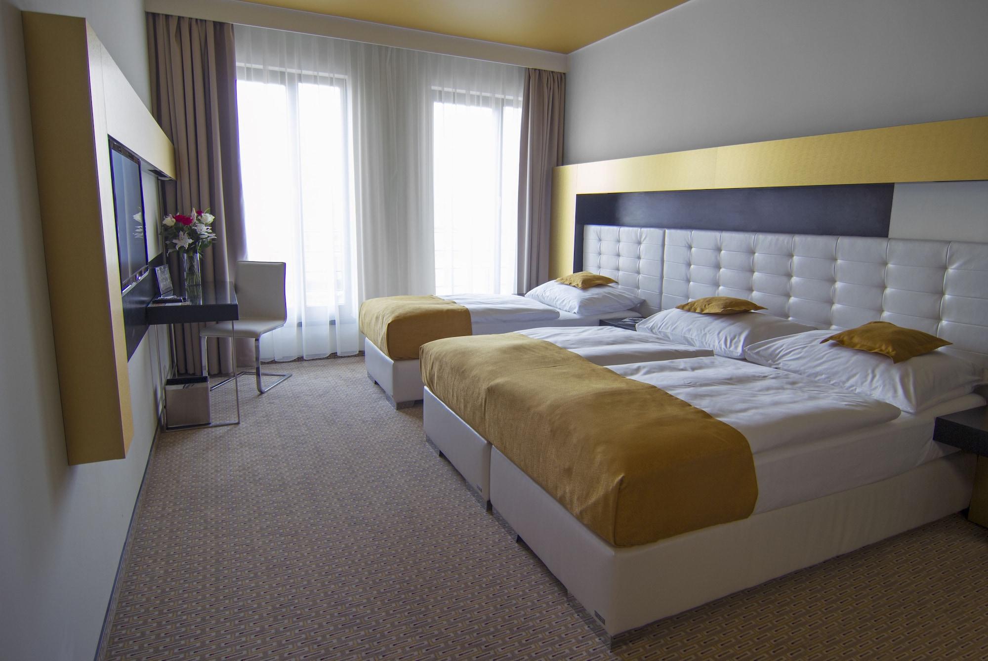Grandior Hotel Praga Esterno foto The photo shows a modern hotel room featuring two beds with white bases and golden bedspreads. The walls are painted in neutral colors with a striking yellow accent along the top. There are large windows dressed with light curtains, allowing natural 