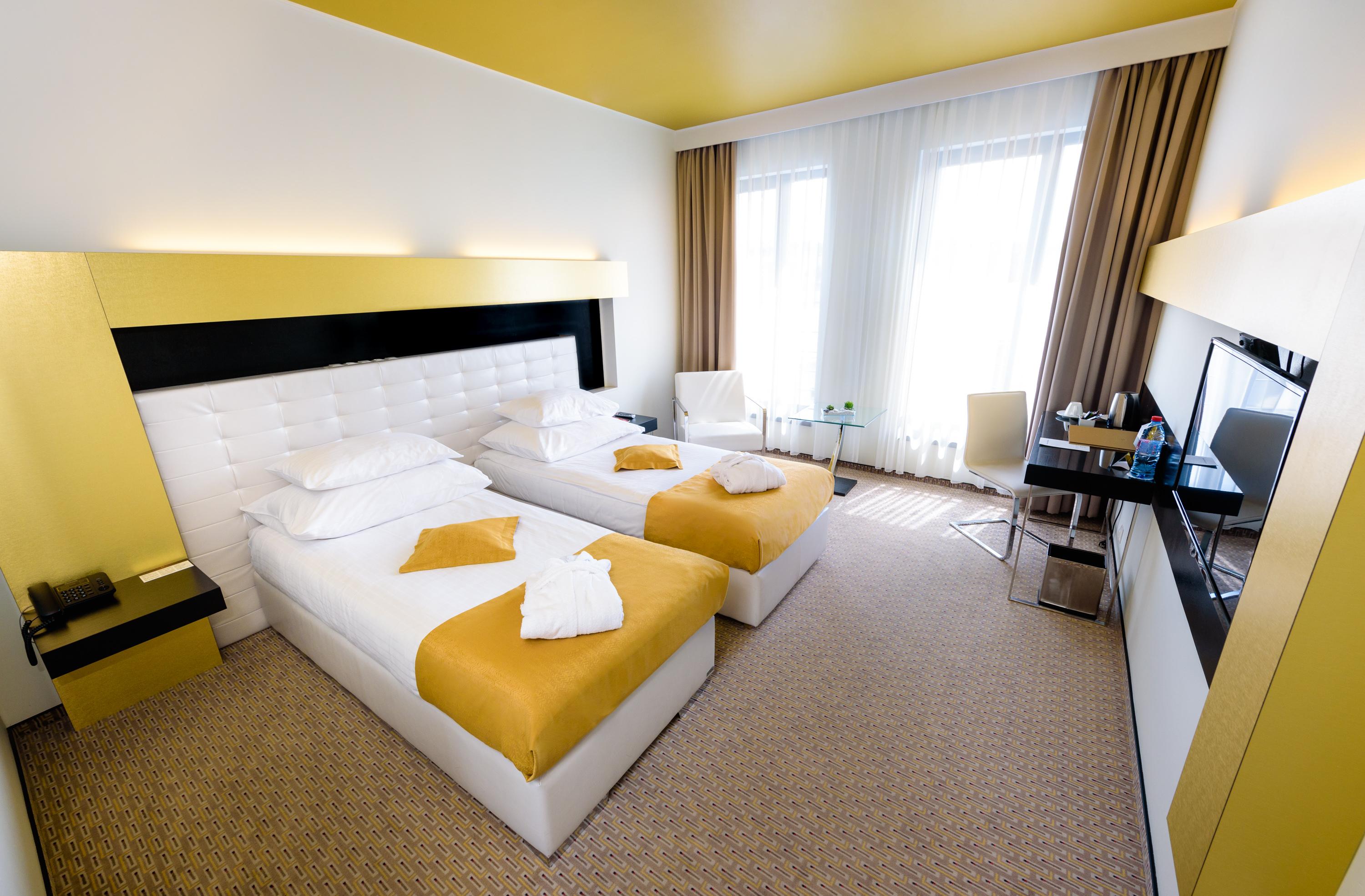 Grandior Hotel Praga Esterno foto The photo shows a modern hotel room featuring two twin beds with white bedding and yellow decorative blankets. The room has a bright, minimalist design, with a yellow ceiling and light-colored walls. Large windows allow natural light to fill the spac